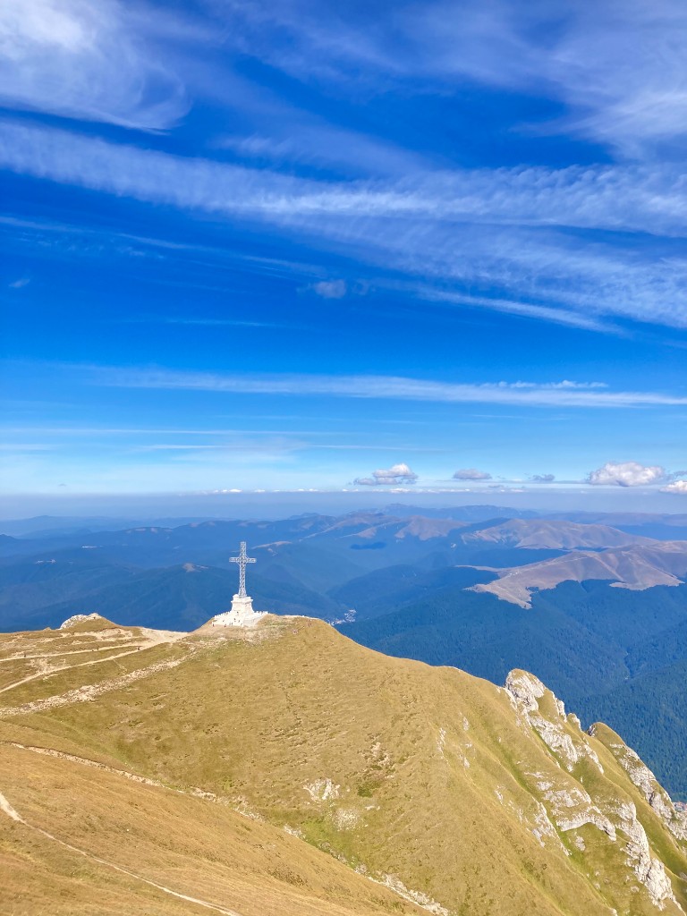 The Caraiman Cross memorial on its stone pedestal on a grassy Bucegi ridge with layered mountain ranges behind